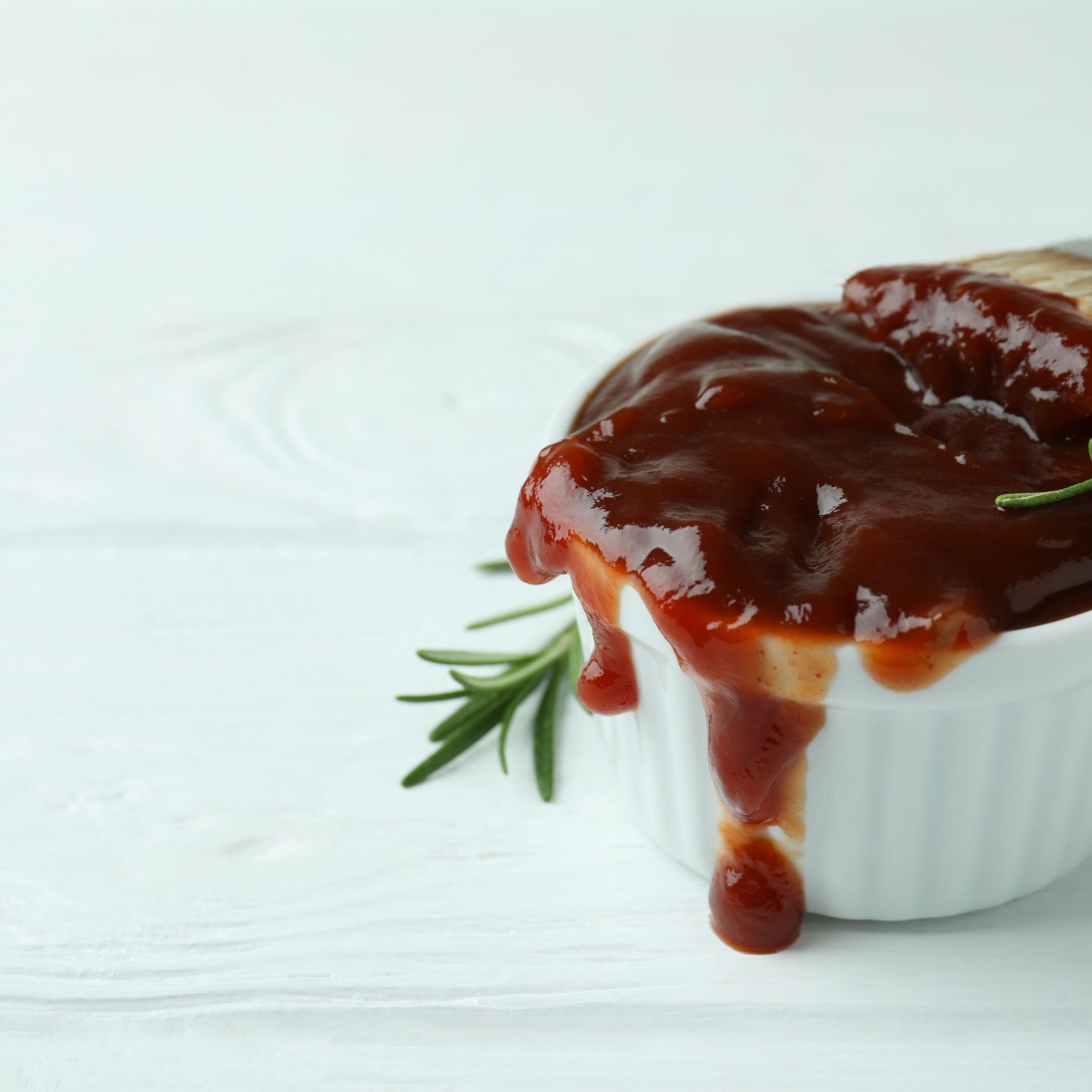 Bowl of barbecue sauce with brush and rosemary on white wooden table
